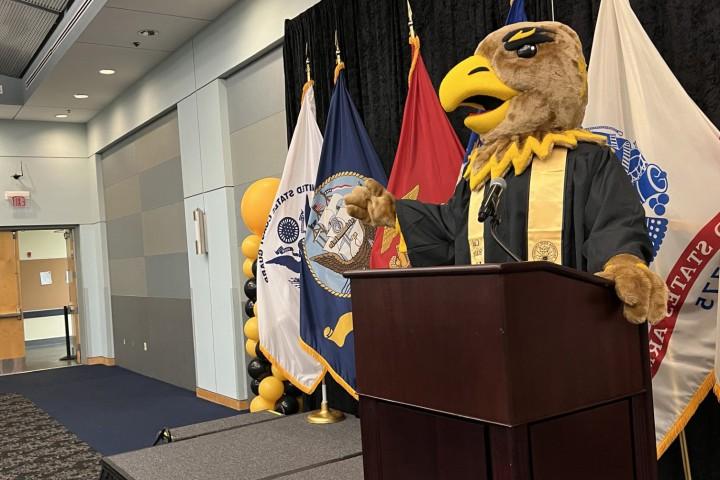Eddie the Golden Eagle wearing a graduation gown, standing at a podium.