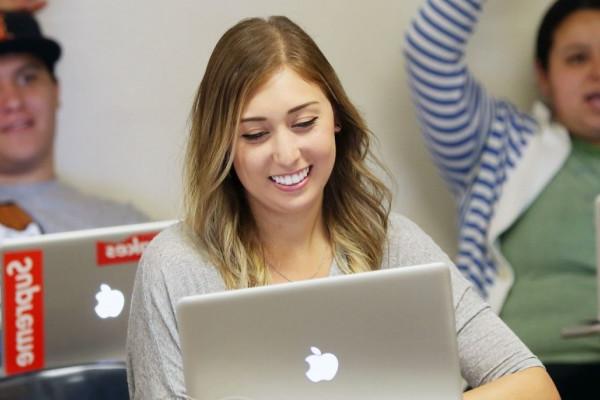 Image of a Caucasian female student smiling in a classroom setting in front of an open laptop. 