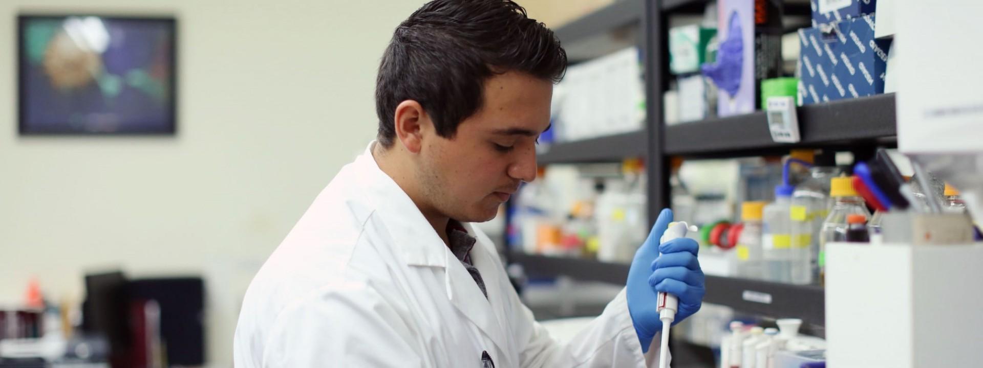 Image of the side profile of a student in a while lab coat and blue gloves using a lab instrument. 