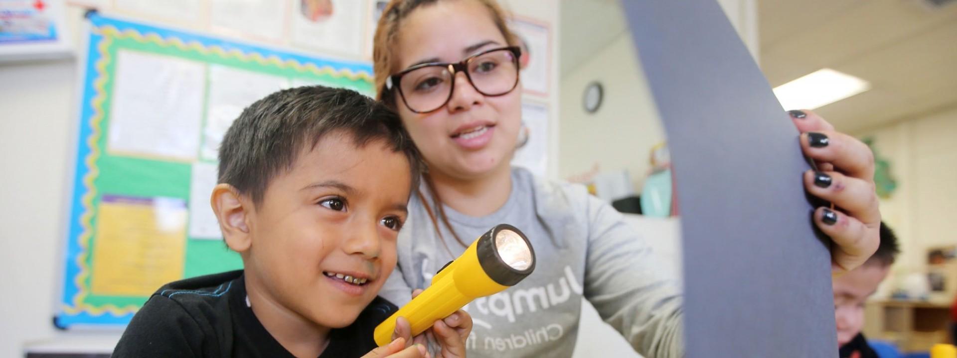 Image of a female student holding up a page for a child who is shining a flashlight on it. 